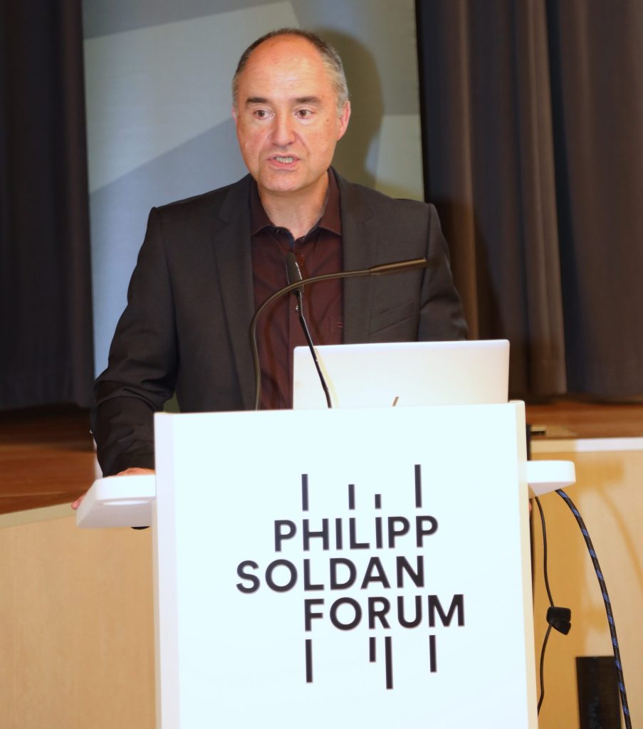 Der Referent Dr. Bernd Breyvogel steht hinter einem Rednerpult und spricht Richtung Plenum. Im Hintergrund sind eine Bühne und Vorhänge zu sehen.