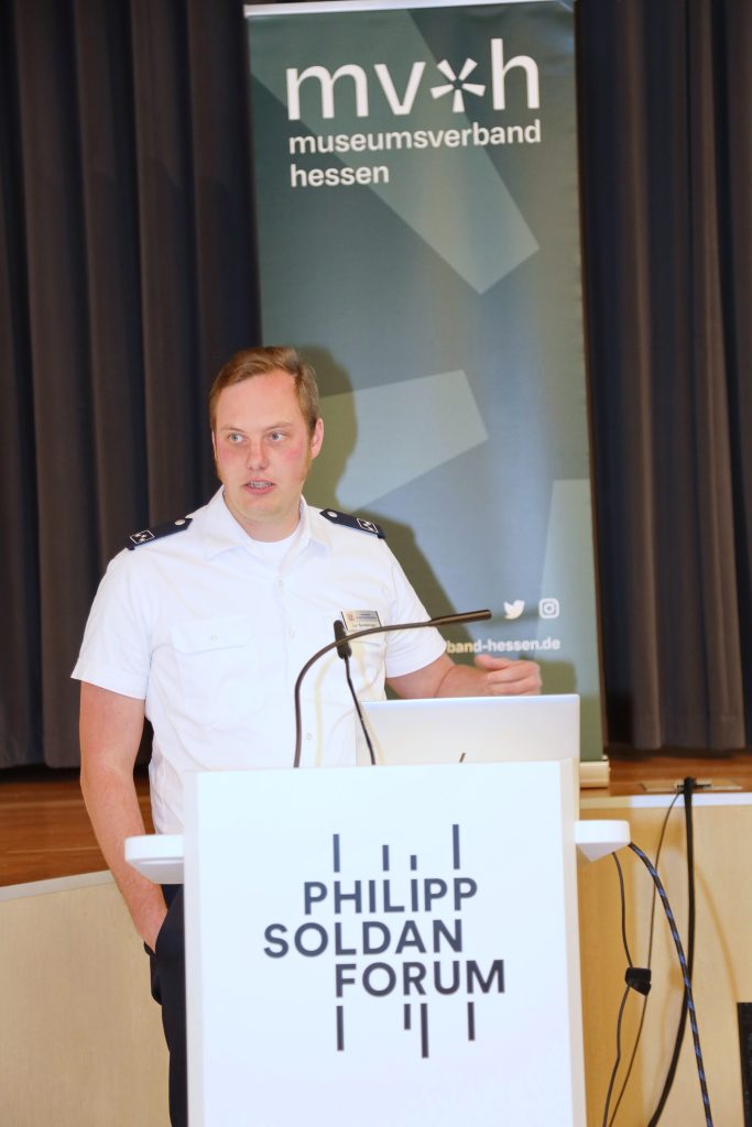 Der Referent Jan Seeberger steht hinter einem Rednerpult und spricht Richtung Plenum. Im Hintergrund sind eine Bühne und Vorhänge sowie das Banner des Museumsverbandes Hessen zu sehen.