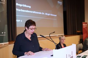 Eine Frau mit Brille spricht am Rednerpult zum Publikum. Auf der Leinwand im Hintergrund ist die Begrüßung durch die Verbandsvorsitzende Dr. Birgit Kümmel angekündigt.