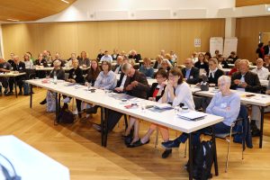 Blick auf die Teilnehmenden des Verbandstags, die an langen Tischen in einem Konferenzraum sitzen und den Vorträgen folgen.