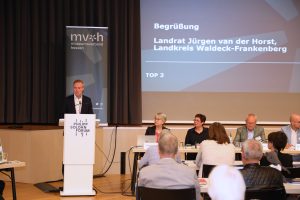 Eine Person steht am Rednerpult, der Blick geht in das Publikum. Auf der Leinwand im Hintergrund ist die Begrüßung durch Landrat Jürgen van der Horst angezeigt.