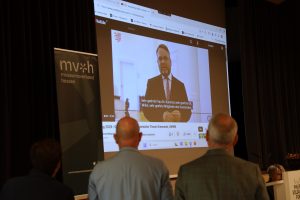 Zwei Personen schauen auf eine Leinwand. Auf der Leinwand ist das digitale Grußwort von Staatsminister Timon Gremmels abgebildet.