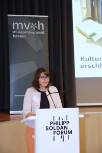 Eine Person mit kurzen braunen haaren und weißer Jacke steht am Rednerpult und spricht ins Publikum. Im Hintergrund ist ein Aufsteller vom Museumsverband Hessen mit Logo zu sehen.
