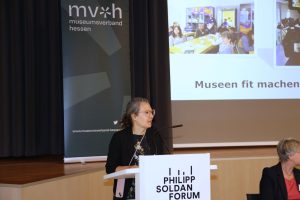 Eine Person mit zurückgebundenen Haaren und dunkler Kleidung steht am Rednerpult und spricht ins Publikum. Im Hintergrund ist ein Aufsteller vom Museumsverband Hessen mit Logo zu sehen. Eine Leinwand ist neben dem Aufsteller zu sehen.