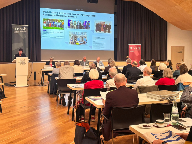 Teilnehmende sitzen an Tischen und verfolgen einen Vortrag. Auf der Leinwand wird eine Präsentation zum Thema Politische Interessenvertretung und kulturpolitische Arbeit angezeigt.